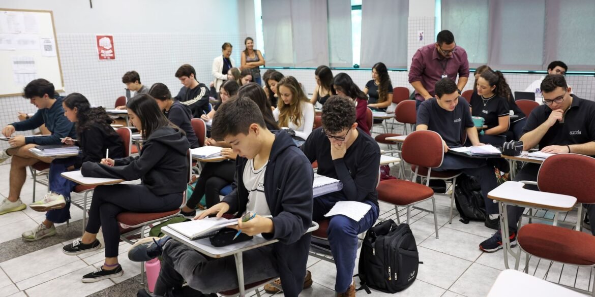 Alunos estudam em sala de aula, acompanhados por professor.