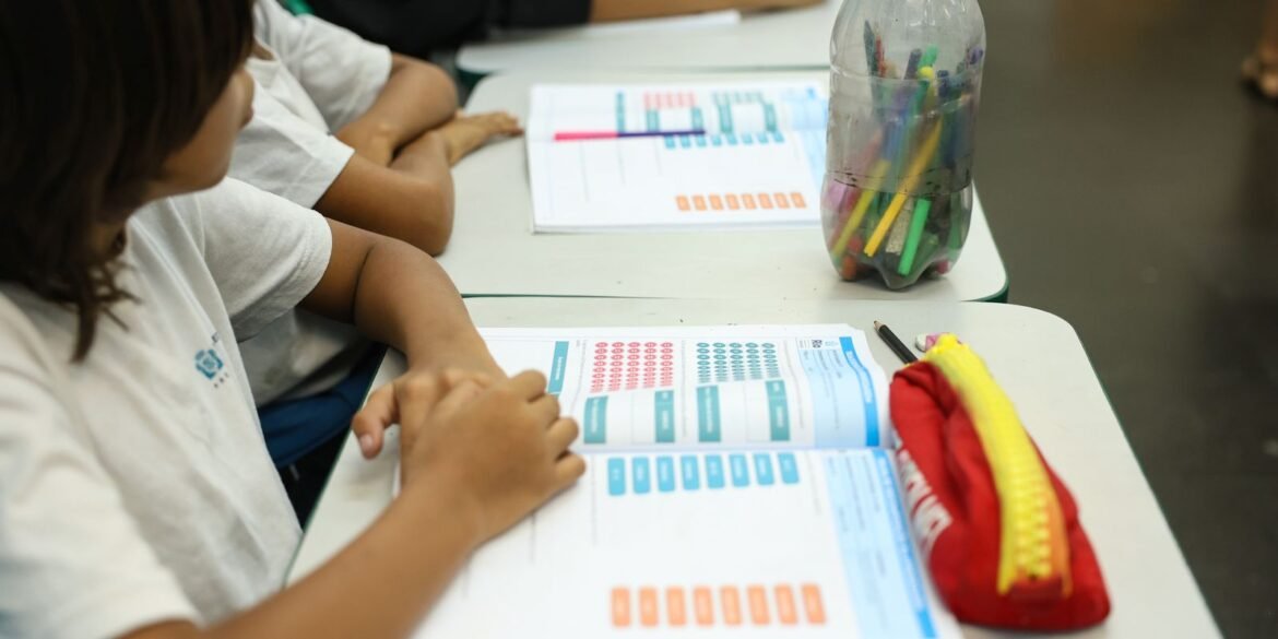 Crianças diante de livros e lápis em mesa escolar.