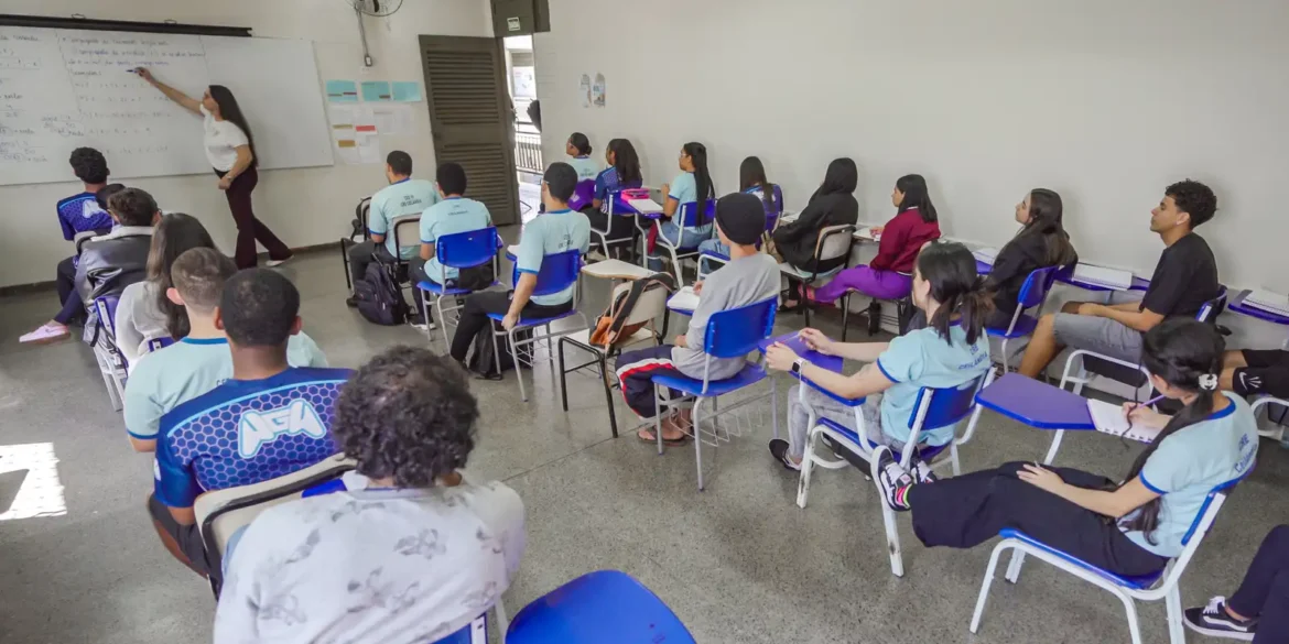 Professora escreve no quadro diante de alunos em sala de aula.