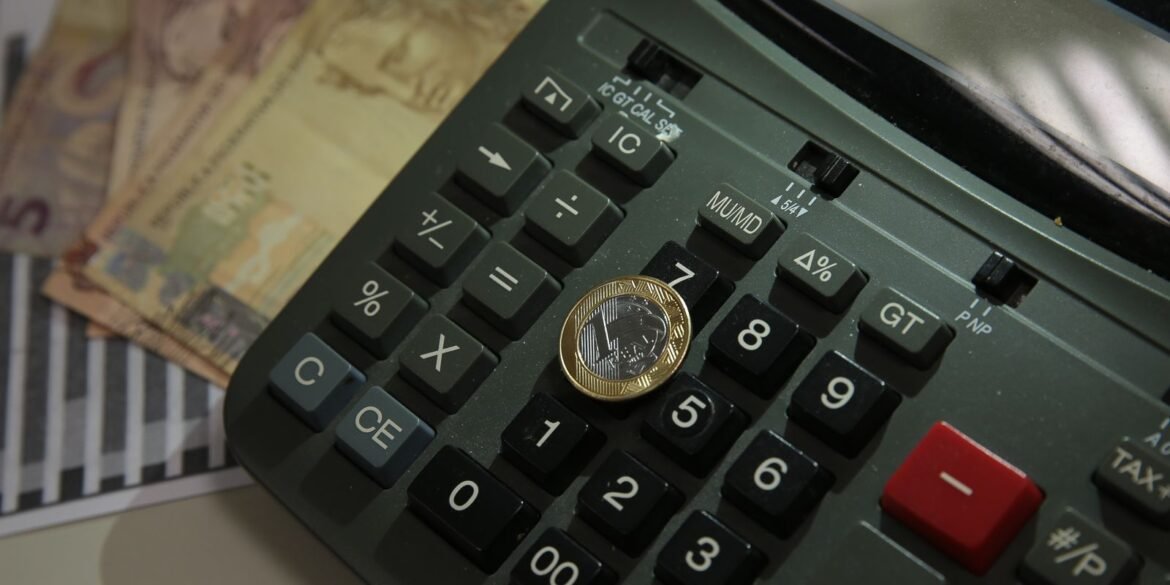 Moeda de um real sobre calculadora, com cédulas ao fundo.